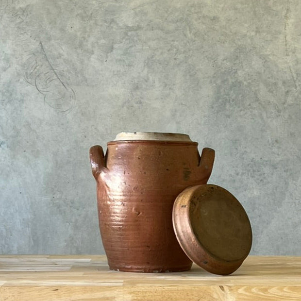 Brown ceramic jar with lid on a wooden surface against a gray wall.