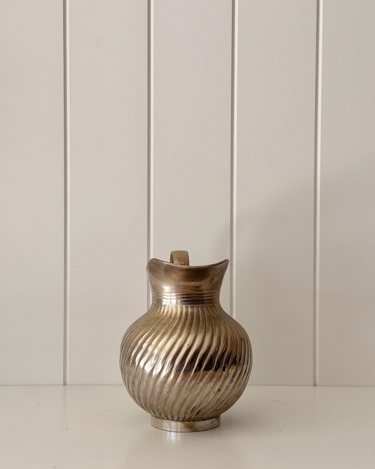 A metallic silver pitcher with a ribbed, fluted texture and a small handle, set against a white paneled background.