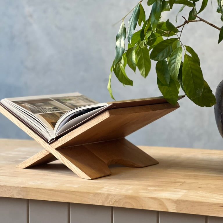 Wooden book stand holding an open book on a wooden surface with a plant in the background.