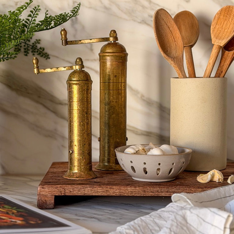 Vintage brass pepper grinder on a wooden stand with kitchen utensils and ingredients.