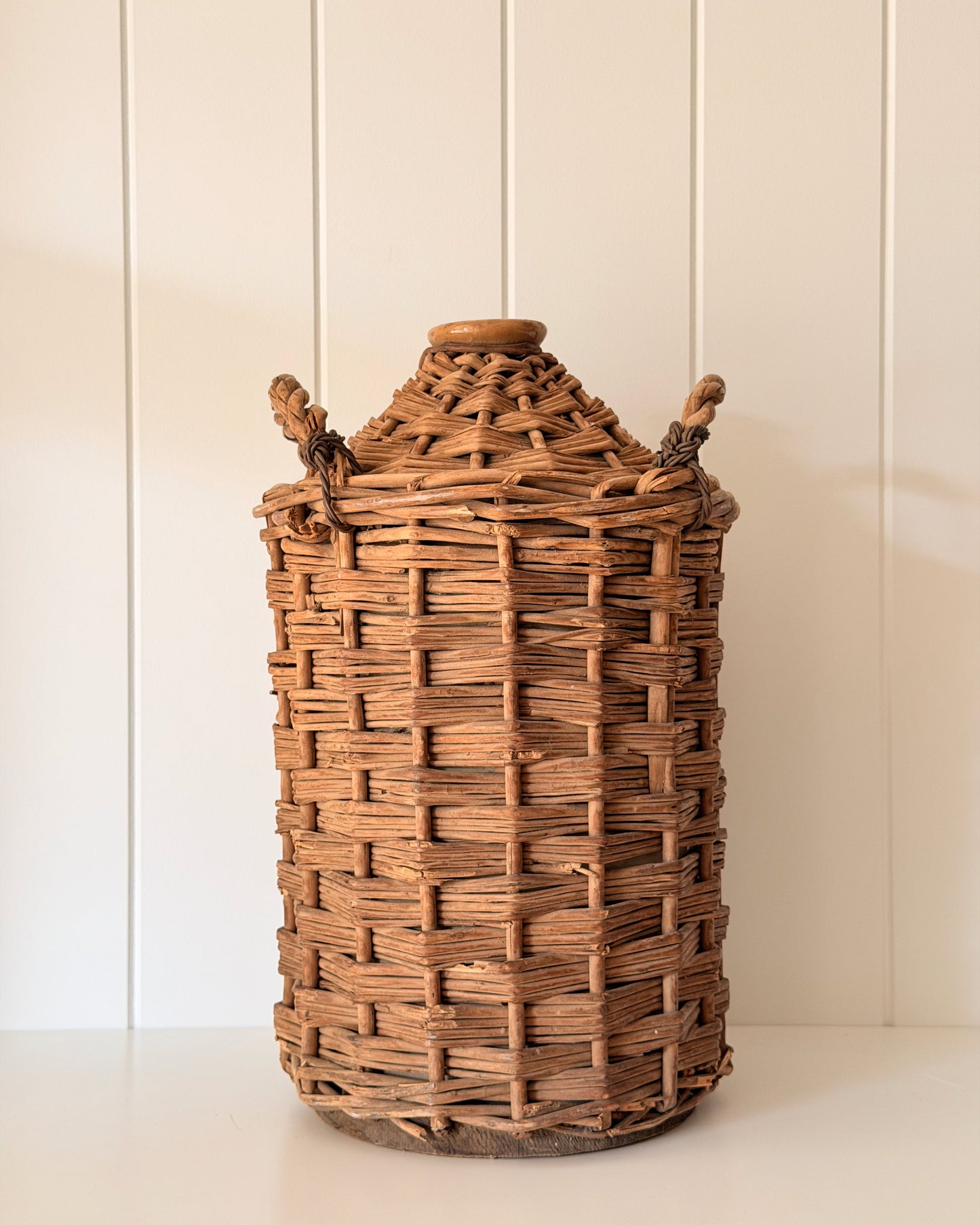 Antique Wicker Demijohn on white tabletop with a panelled wall.