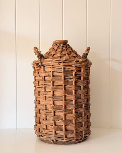 Antique Wicker Demijohn on white tabletop with a panelled wall.