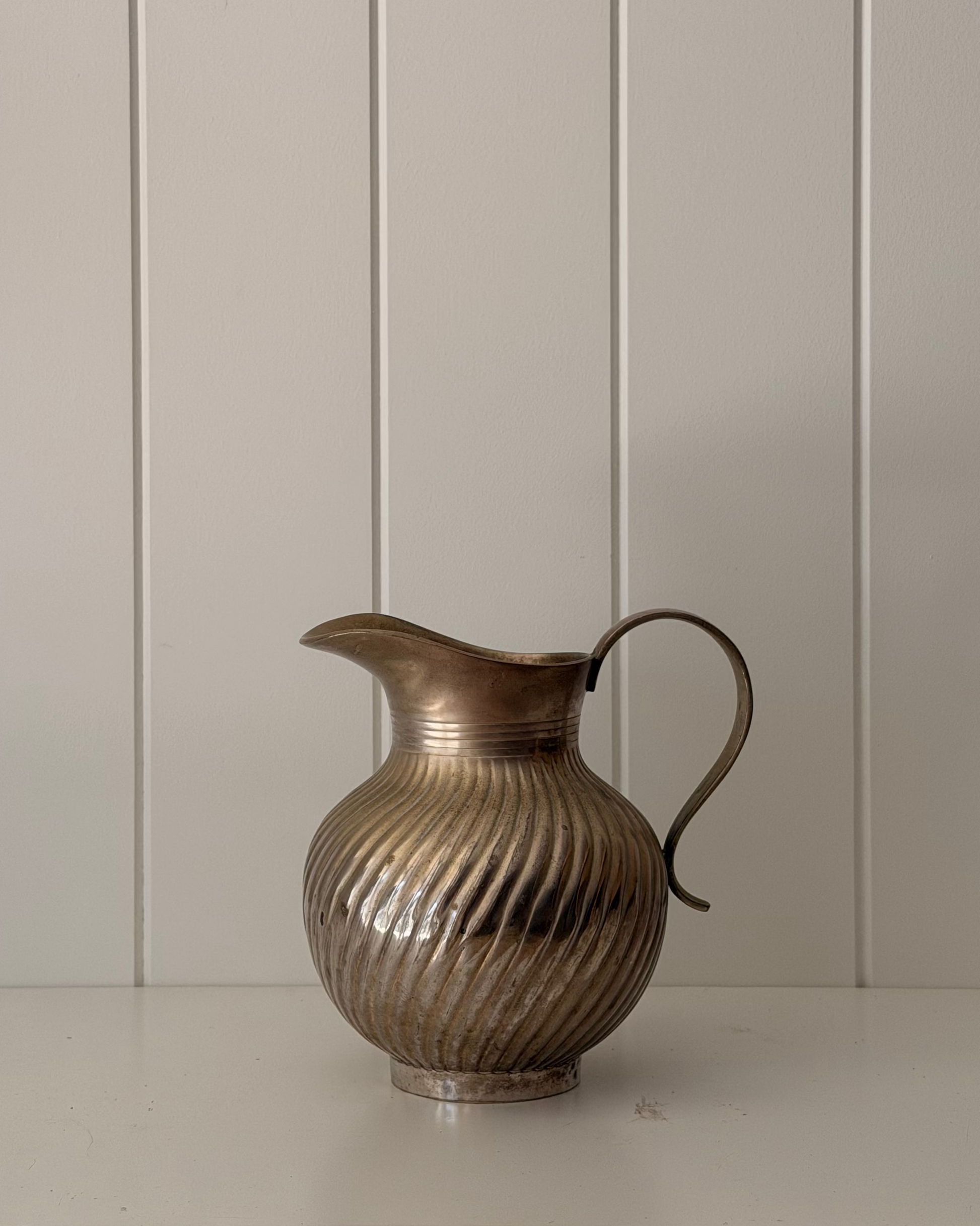 A vintage-style metallic pitcher with a ribbed, vertically fluted body and a curved handle, sitting on a white shelf against a white paneled wall.
