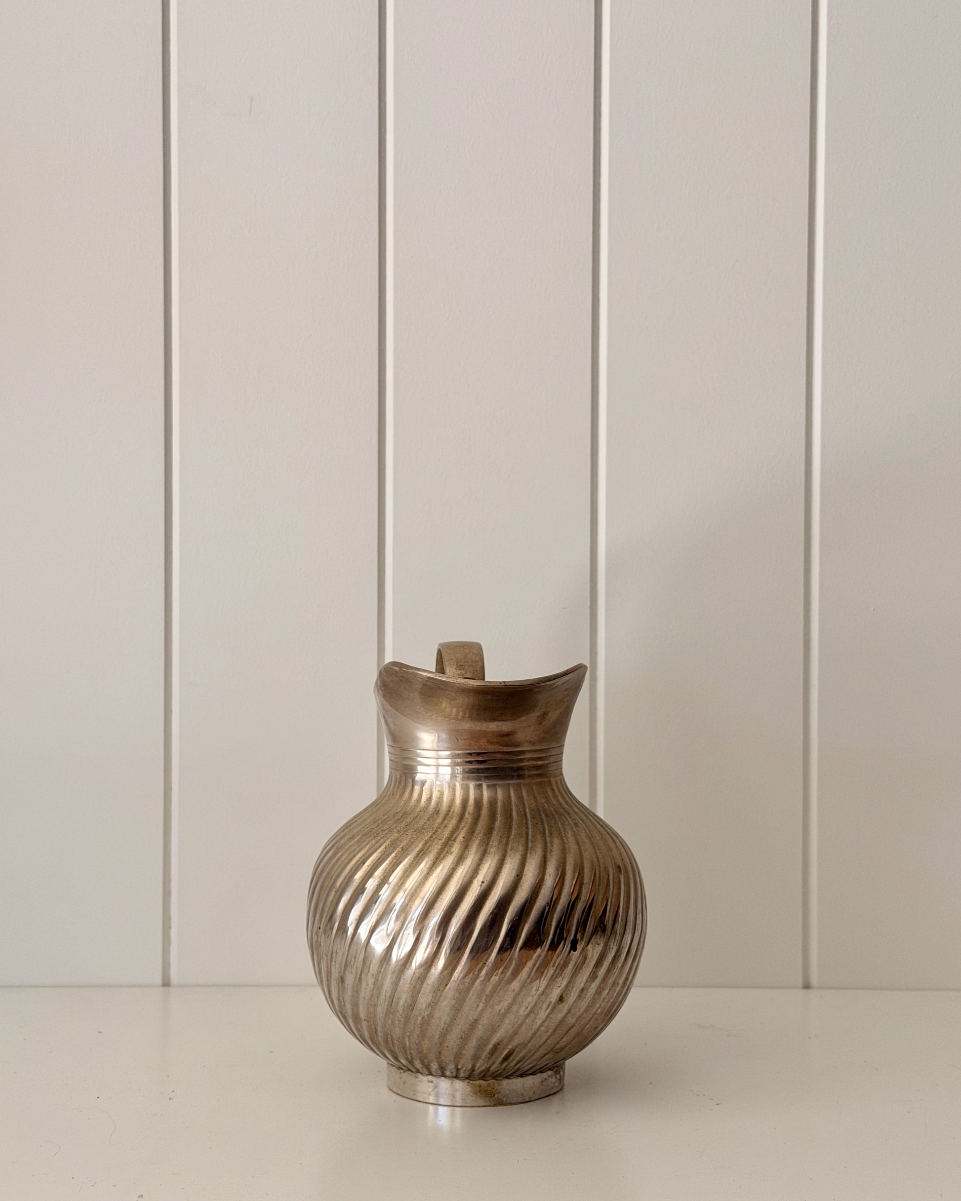 A metallic silver pitcher with a ribbed, fluted texture and a small handle, set against a white paneled background.