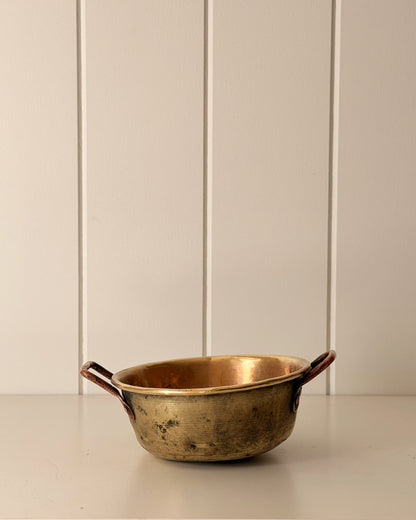 A vintage brass-colored metal bowl with dark brown handles, set against a cream-colored paneled wall.