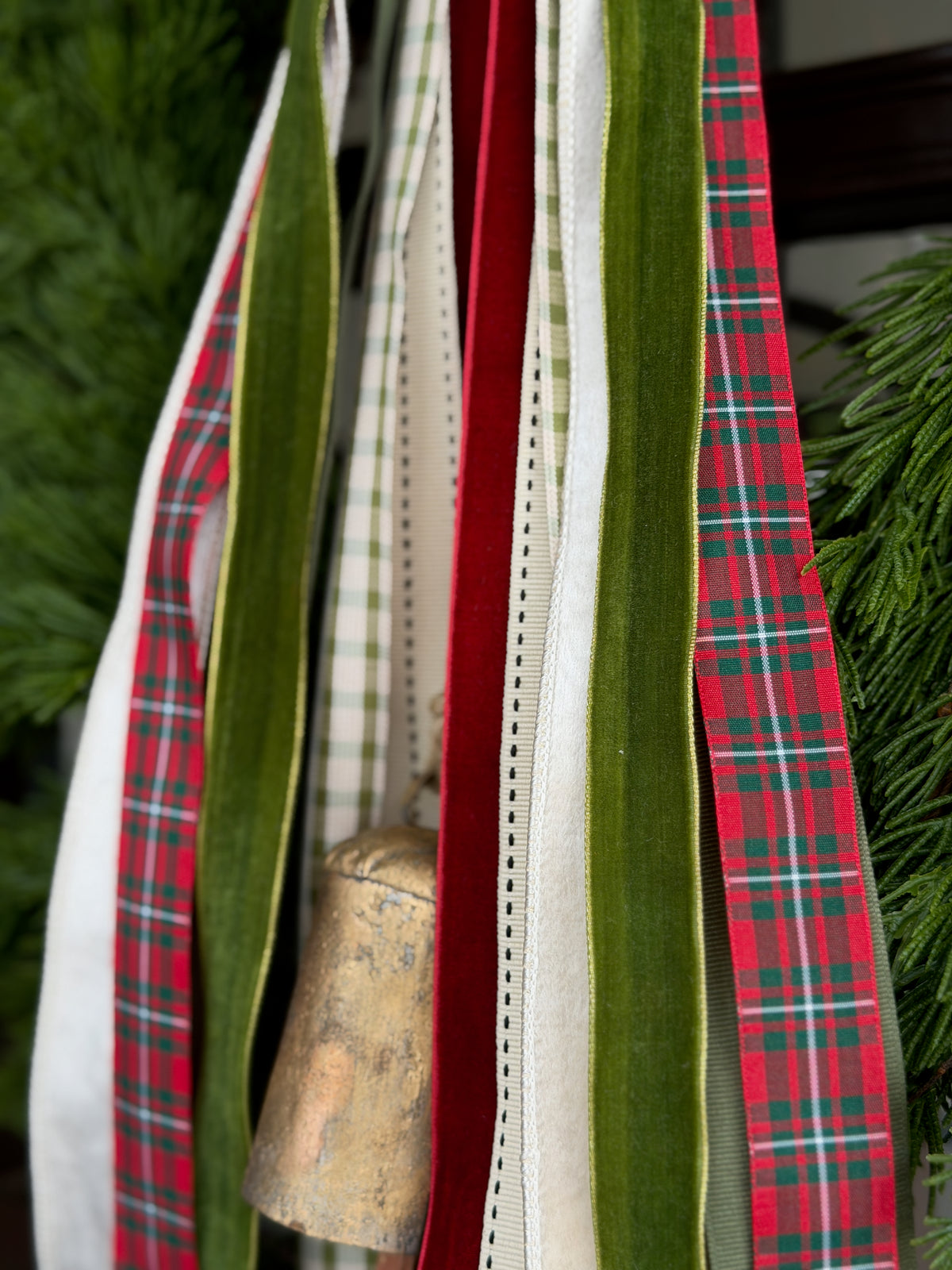 A close-up of hanging Christmas ribbons in festive colors, including red plaid, green velvet, cream stripes, and a red-green tartan ribbon, with evergreen branches and a small metallic ornament visible in the background.