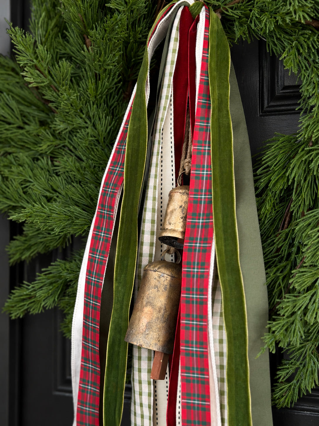 A festive holiday wreath decoration featuring a large bronze bell hanging from layered ribbons. The ribbons include a wide green velvet ribbon, a red and green plaid ribbon, a white and green gingham ribbon, and a solid red ribbon, all set against a dark door and green pine branches.